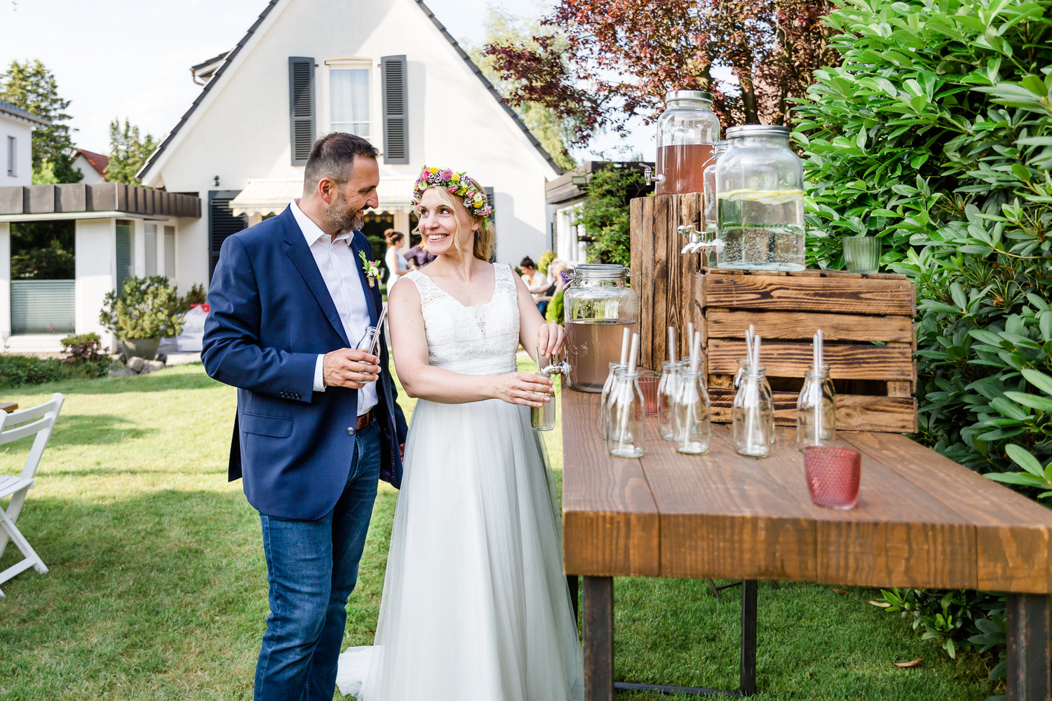 Lächelndes Brautpaar genießt Getränke bei einer Hochzeit im Garten mit Getränkestation und Glastrinkhalmen.