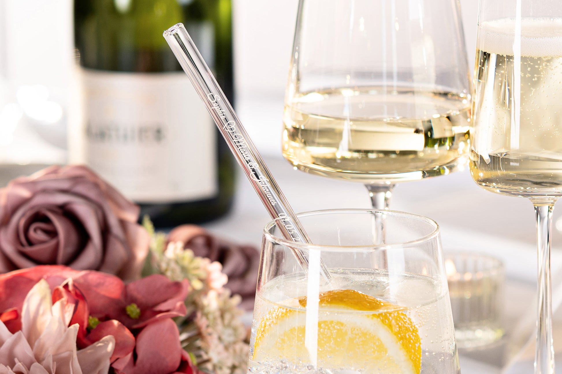 Personalisierter Glasstrohhalm mit Gravur in Getränk, umgeben von Blumen und Champagnerflasche.